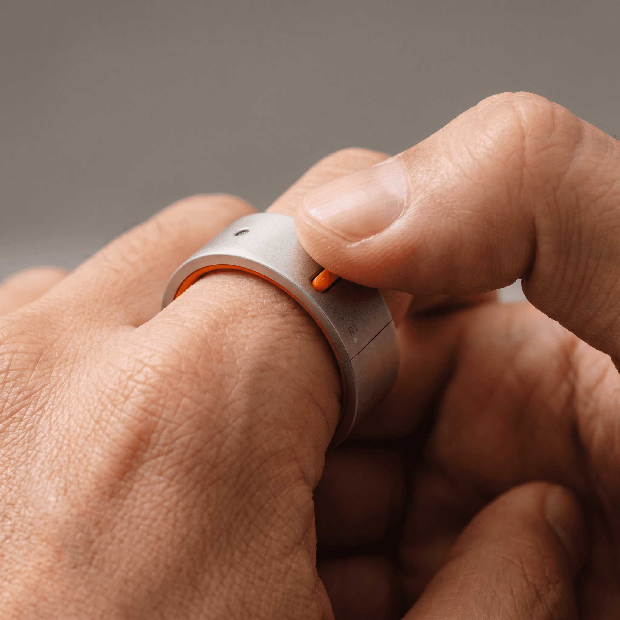 A thumb pressing the recessed orange button on the Rhyme Devices R1 ring worn on the index finger
